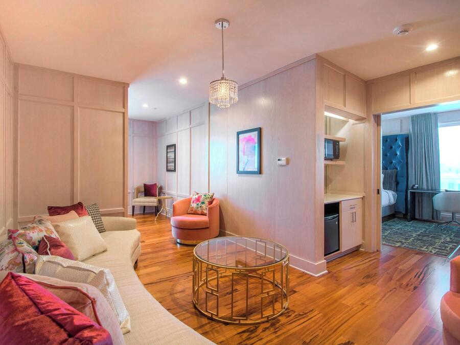 Een moderne woonkamer met een bank, fauteuil en een rond bijzettafeltje. Er is een kitchenette met een magnetron en een kleine koelkast. De kamer heeft houten vloeren en een kroonluchter.