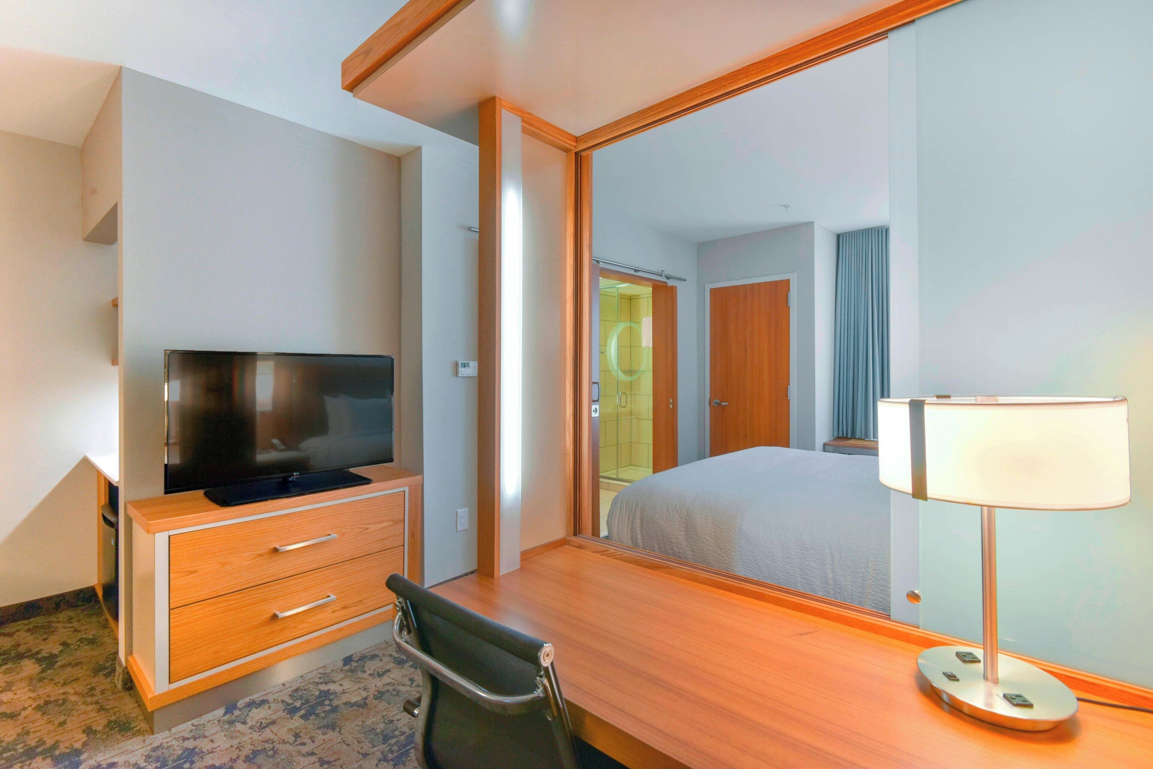 Una habitación de hotel con un escritorio de madera, televisión, espejo y lámpara de noche.