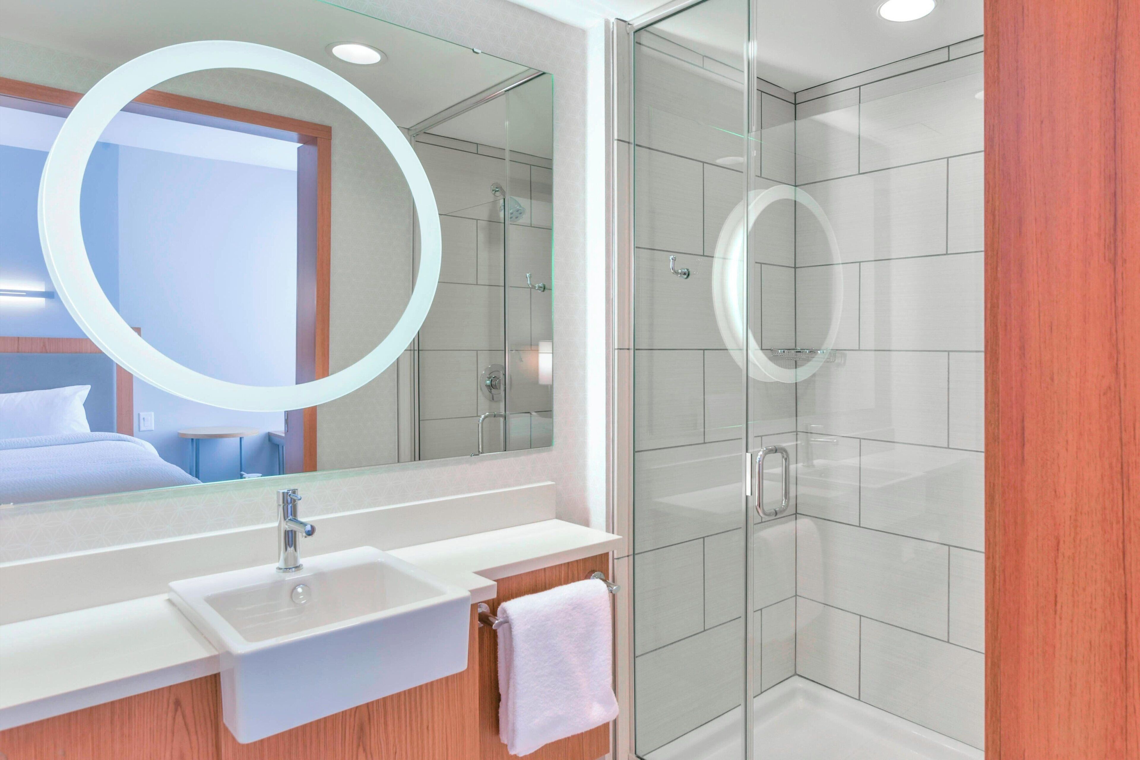 Ein modernes Badezimmer mit einer glasumschlossenen Dusche, einem großen Spiegel mit rundem Rahmen und einem weißen Waschbecken mit einem darüber hängenden Handtuch.