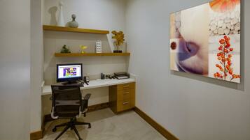 A home office with a desk, computer, and chair. There are shelves with decorative items and a painting on the wall.