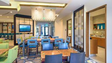 Un espace de restauration d’hôtel avec une grande table et des chaises, un téléviseur et un lustre moderne.