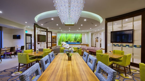 A hotel lobby with a large wooden conference table surrounded by green and blue patterned chairs, a chandelier, and a green-themed backdrop with a television.