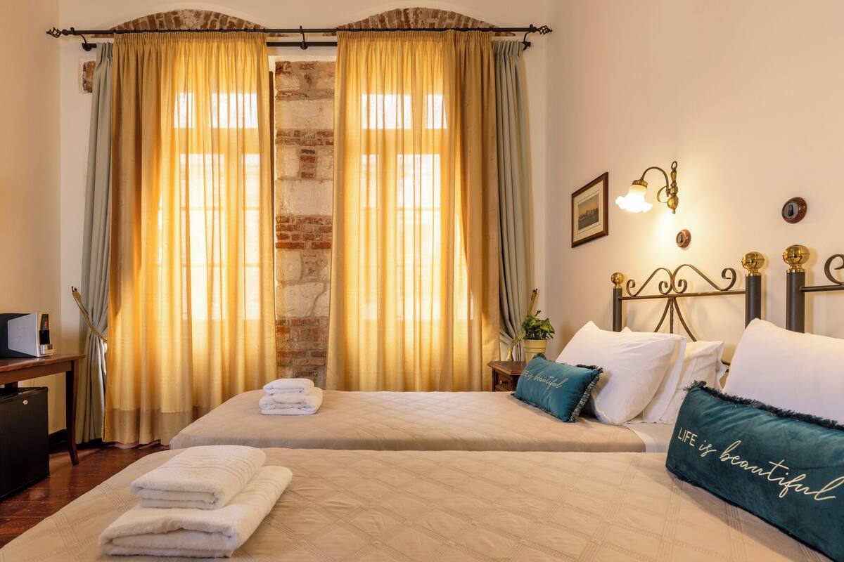 A double bed with a beige quilt, a bedside table with a lamp, and a window with curtains.