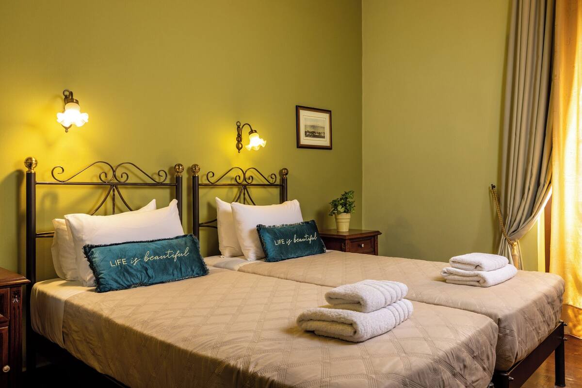 A double bed with two pillows, a bedside table, a framed picture, and a window with curtains.