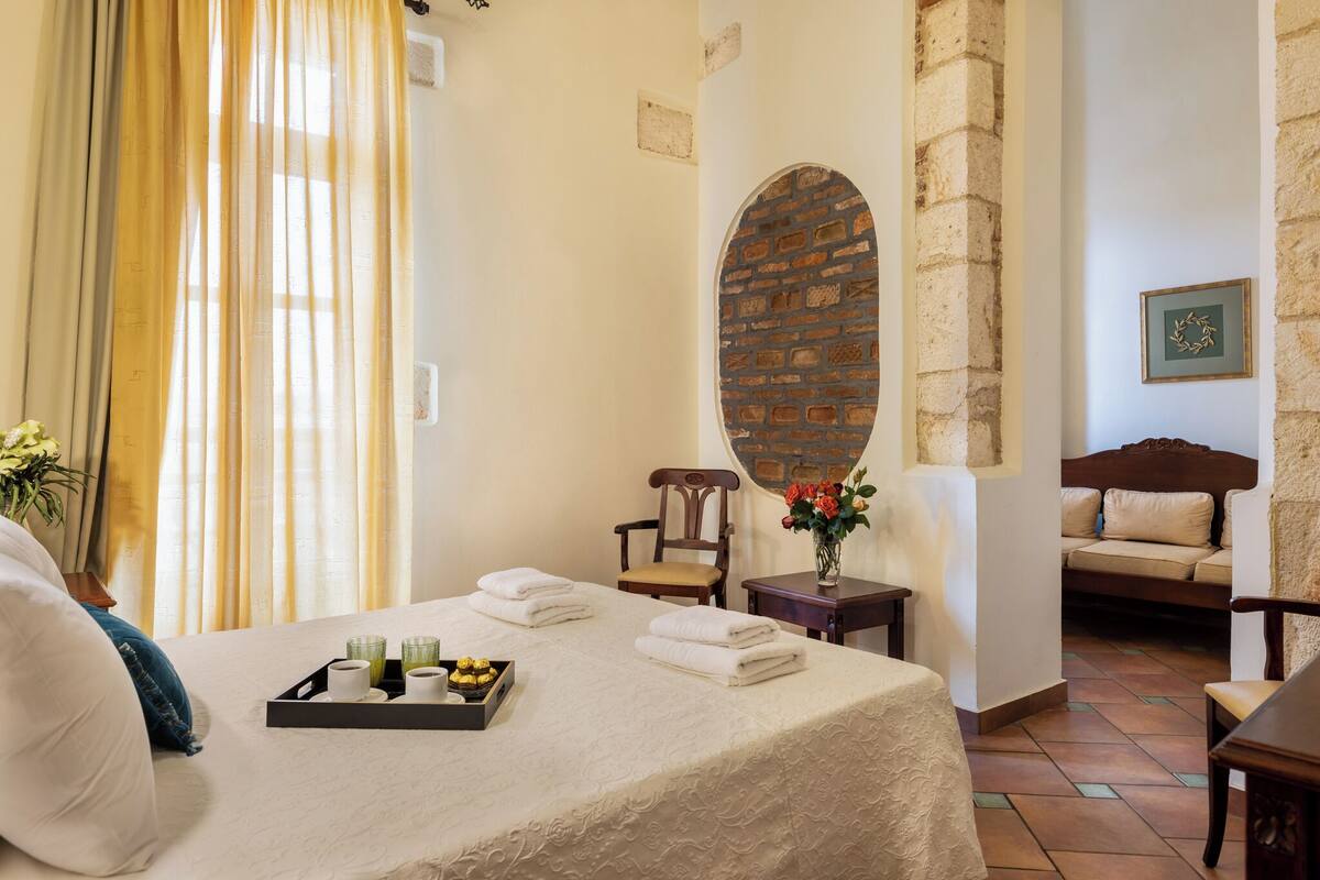 A hotel room with a bed, a small table with a tray of toiletries, a chair, a sofa, and a decorative brick wall.