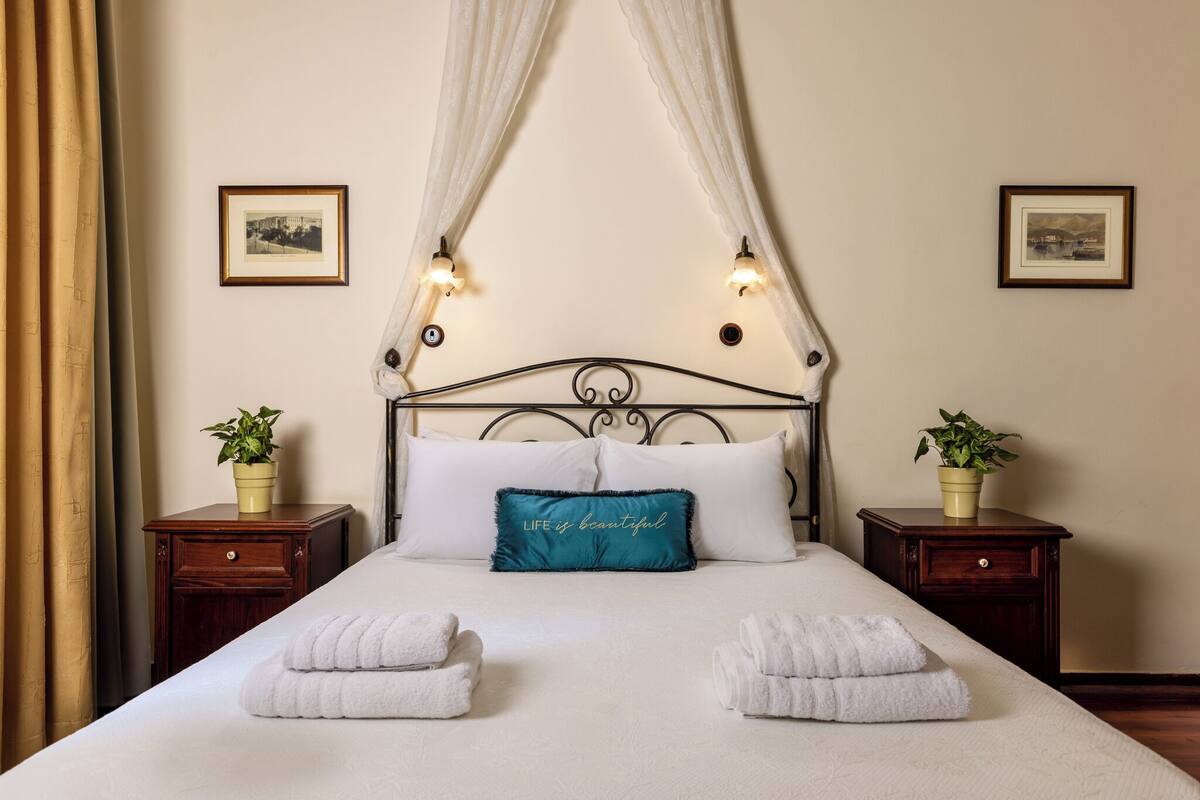 A neatly made bed with white linens, a teal pillow with the text "LIFE is beautiful", and two bedside tables with potted plants.