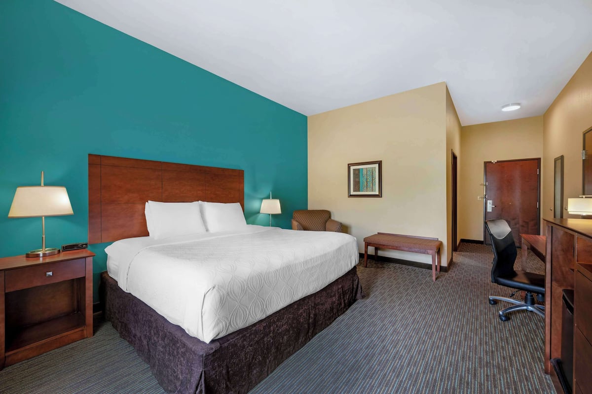 A hotel room with a large bed, a desk, a chair, a lamp, a bench, and a door.