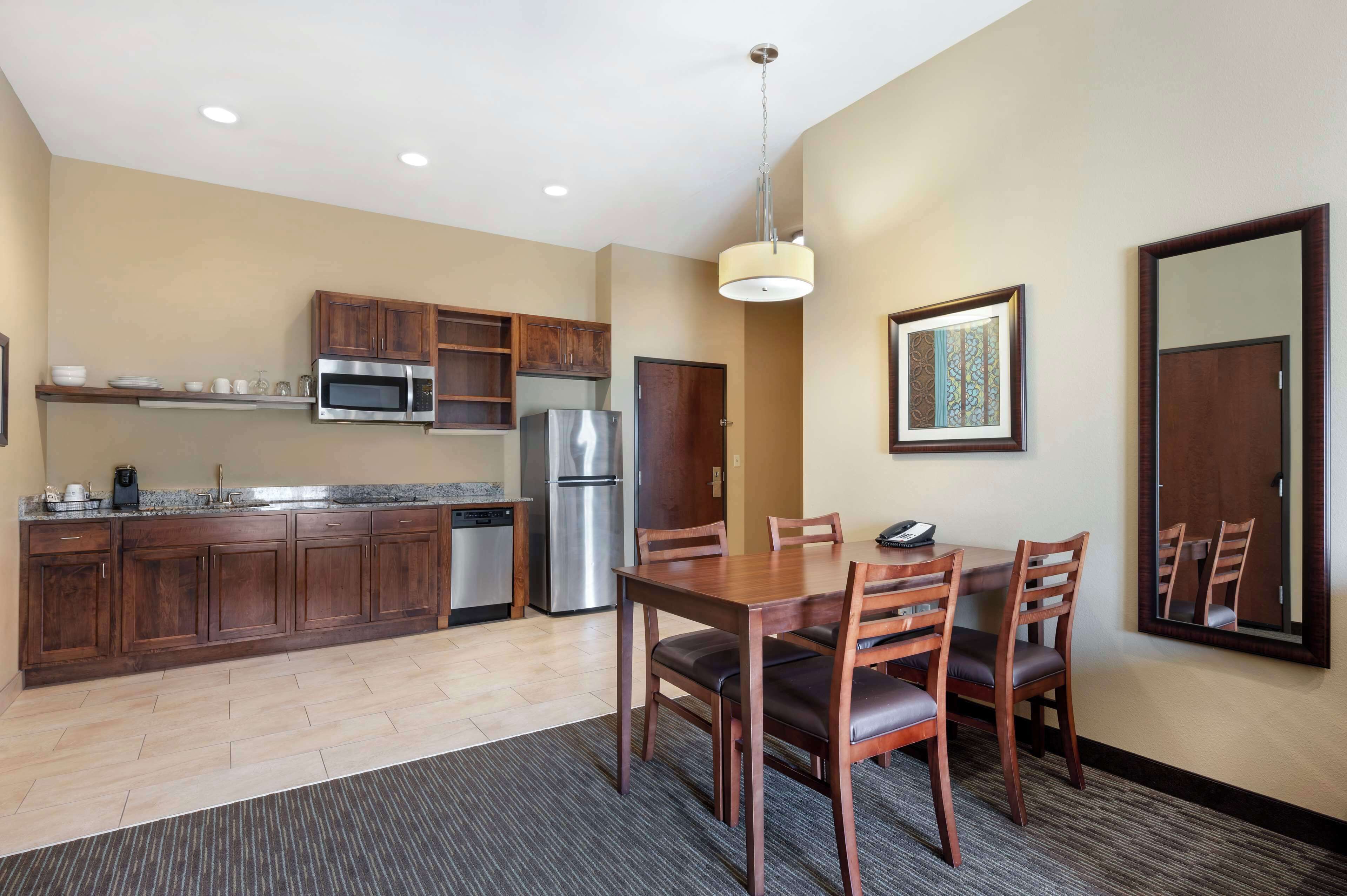 A modern kitchen with wooden cabinets, a stainless steel refrigerator, and a microwave. There is a dining table with chairs and a hanging light fixture above it.