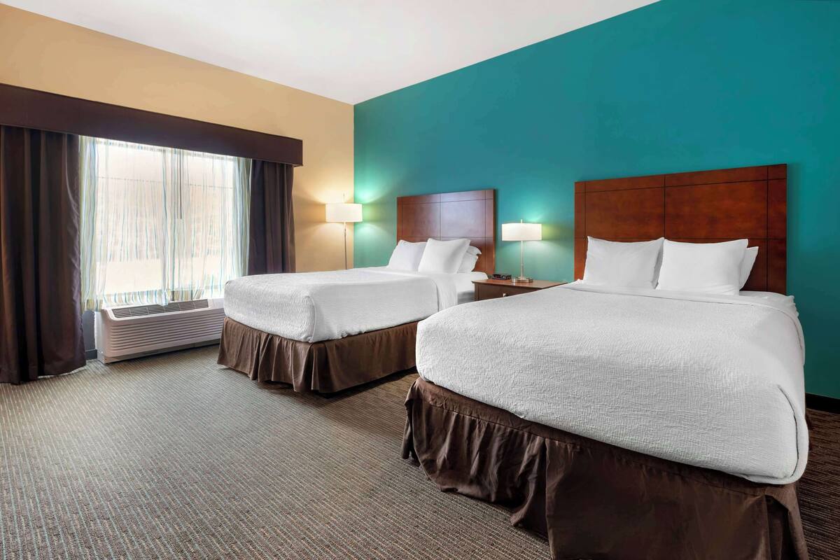 A hotel room with two beds, a large window with curtains, an air conditioning unit, and two bedside lamps.
