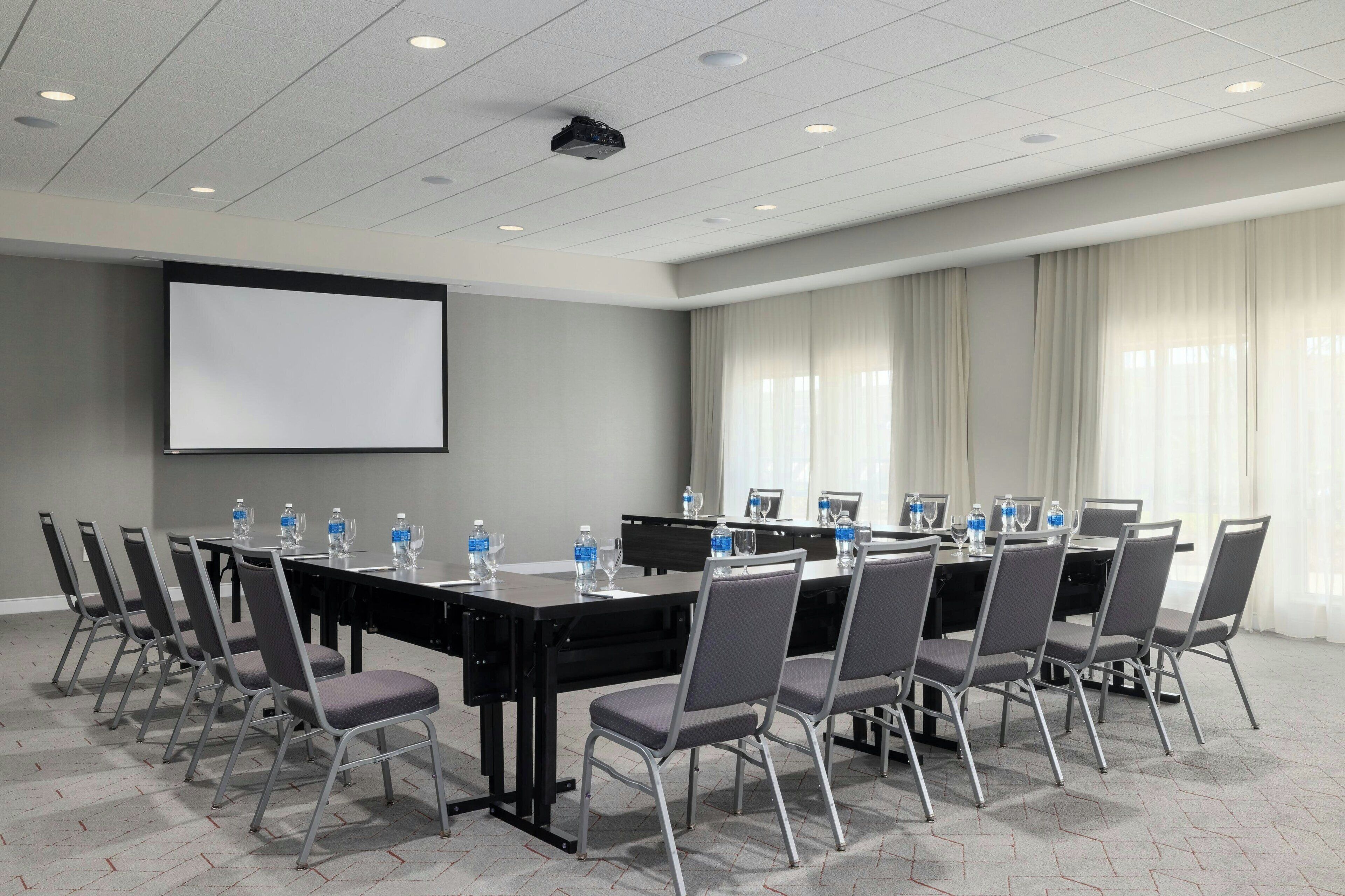 Un salón de conferencias con una larga mesa, sillas, una pantalla de proyector y un amplio ventanal con cortinas.