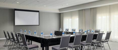 Un salón de conferencias con una larga mesa, sillas, una pantalla de proyector y un amplio ventanal con cortinas.