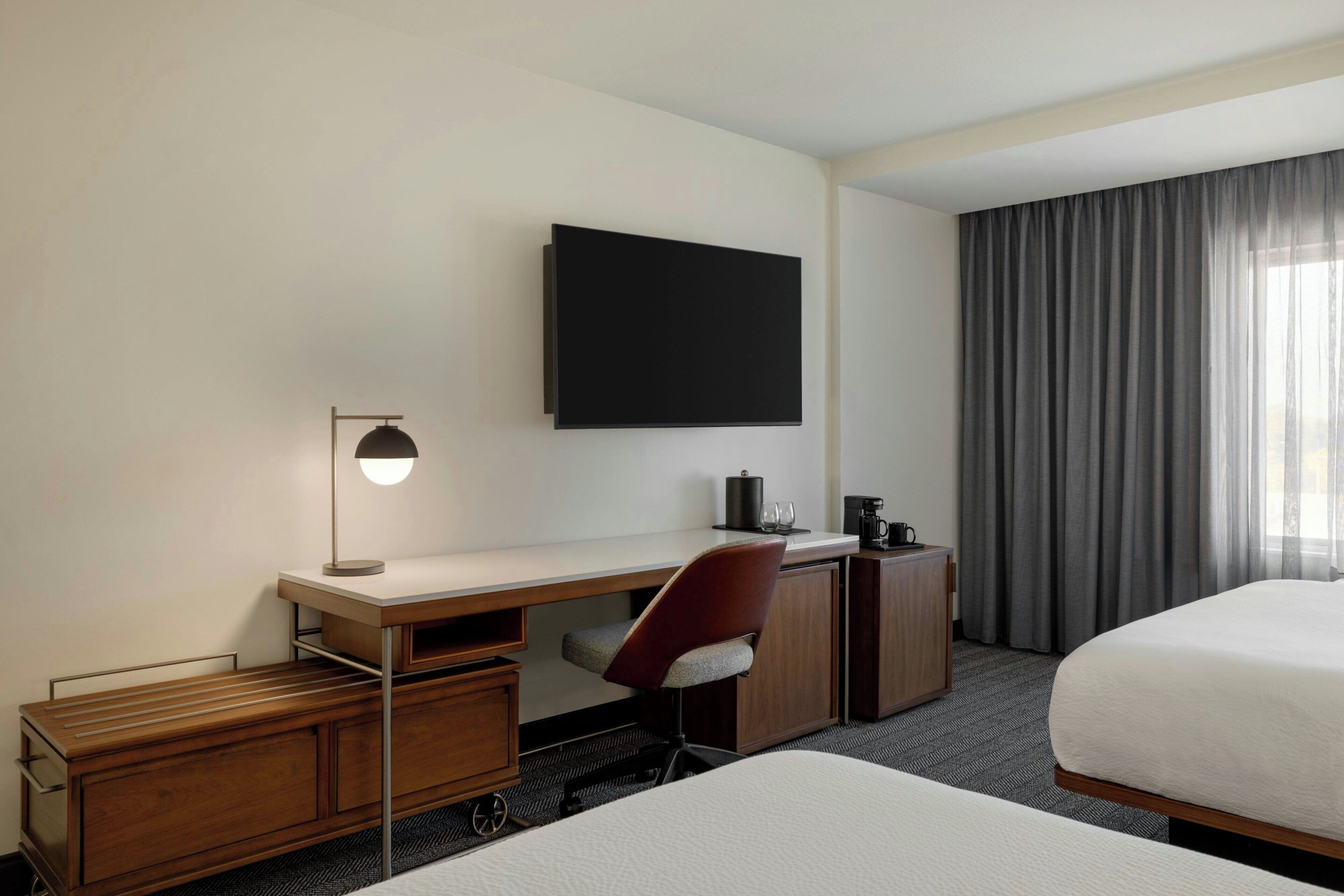 Habitación de hotel con escritorio, una silla, cama, televisor montado en la pared y ventana con cortinas.