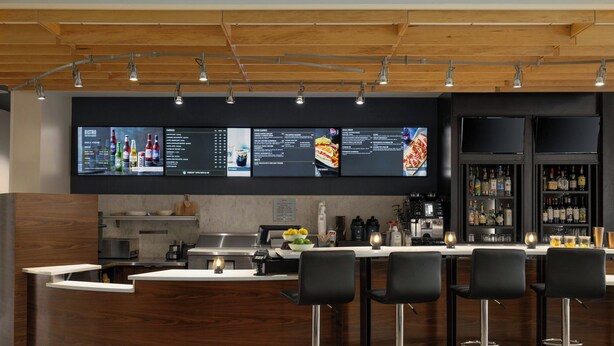 A modern bar area with a wooden ceiling, a digital menu display, and bar stools. - Courtyard by Marriott Starkville MSU at The Mill (Starkville)