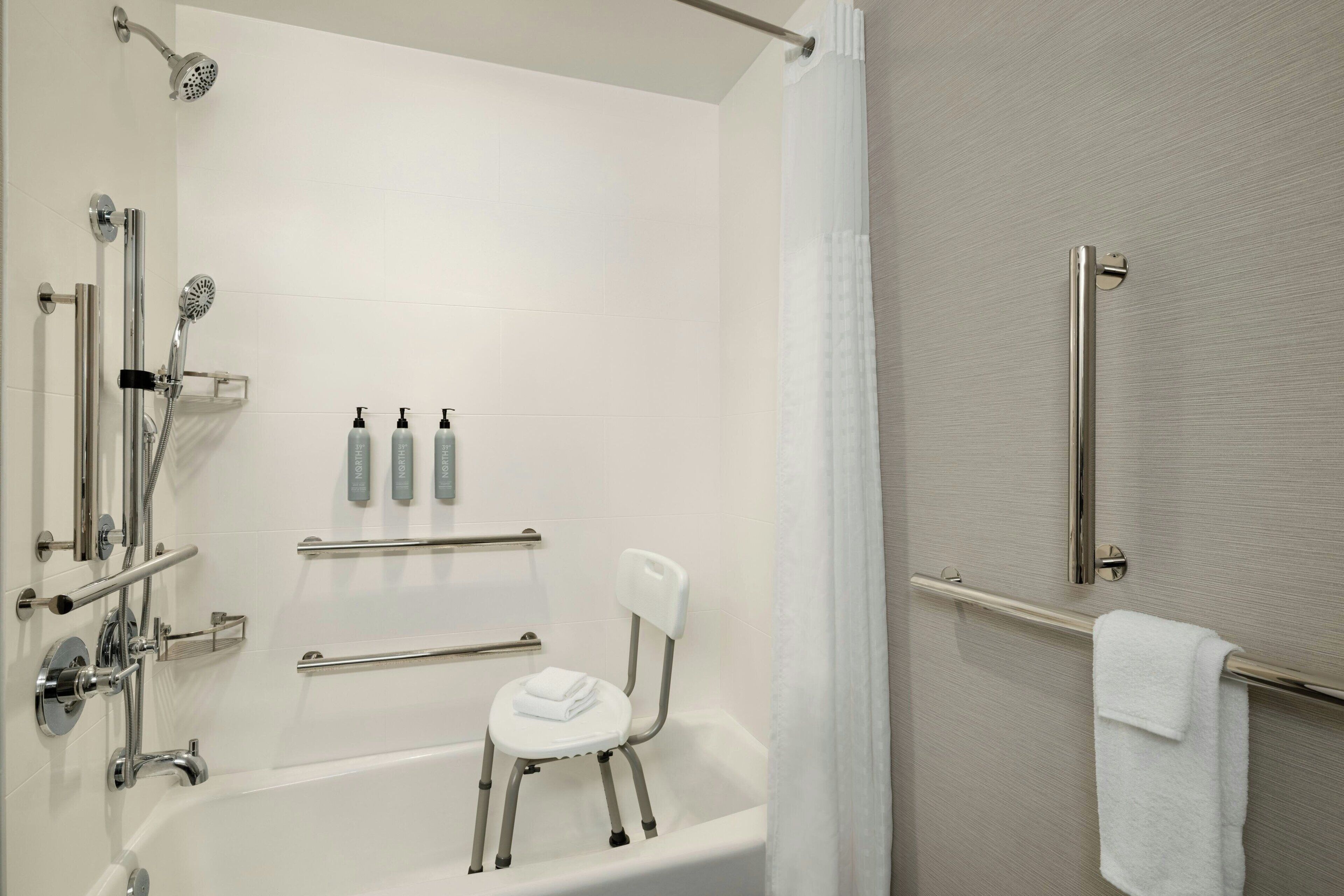 Un baño con bañera, ducha, barandilla, barra de apoyo, una silla blanca y un perchero.