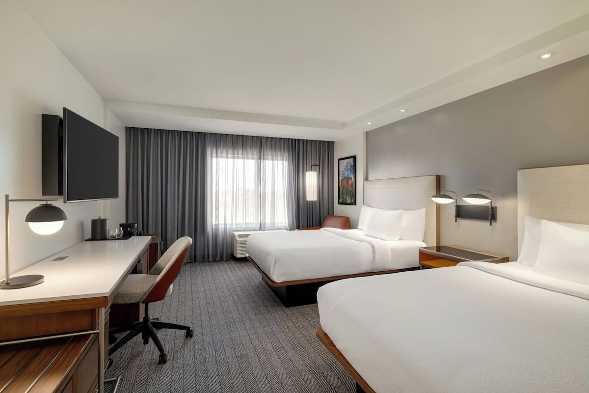 Une chambre d’hôtel avec deux lits, un bureau, une chaise, une télévision et une fenêtre avec des rideaux.