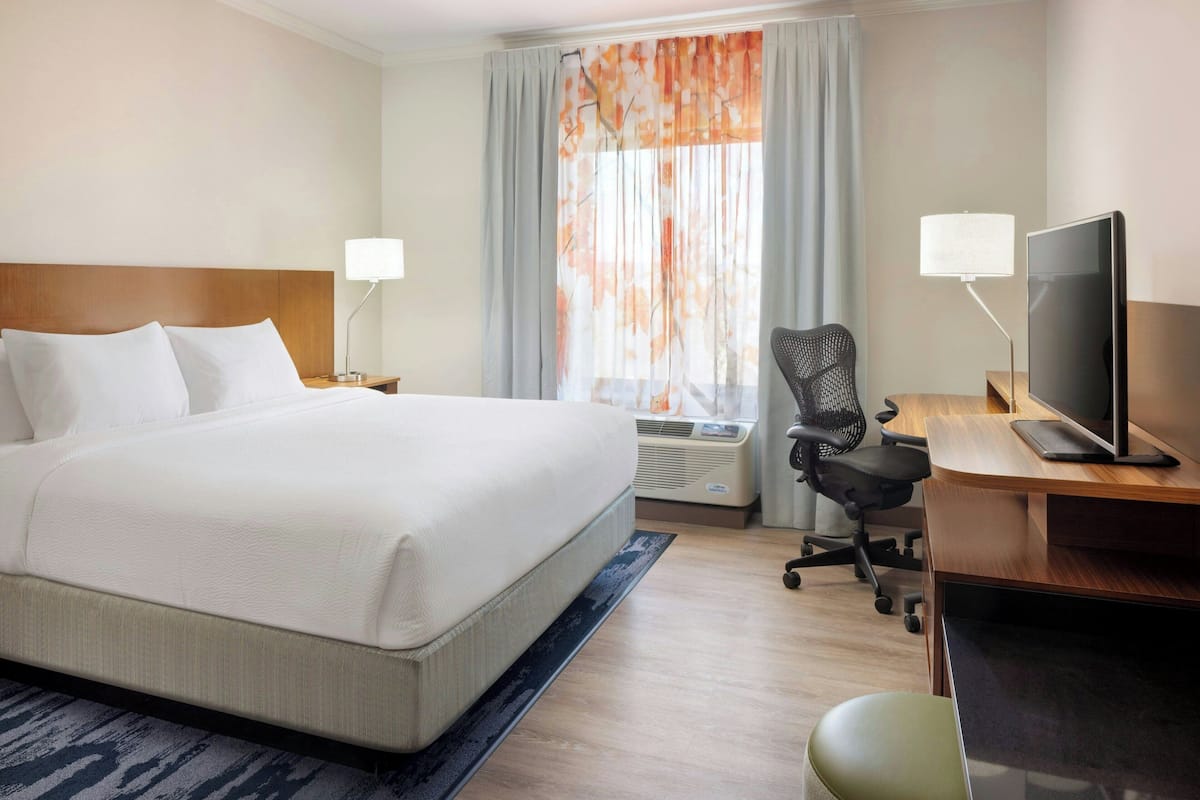 A hotel room with a large bed, a desk with a computer, a chair, and a window with curtains.