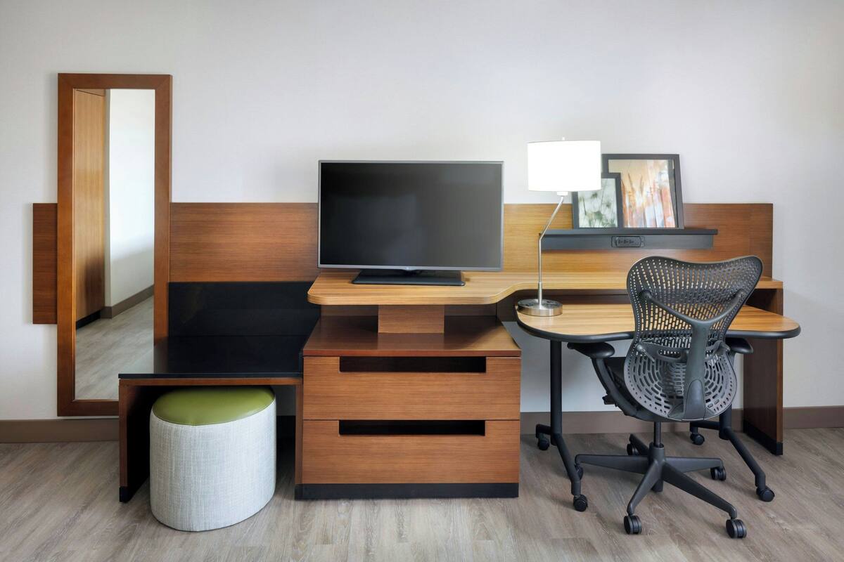 A modern hotel room with a wooden desk, a flat-screen TV, a black office chair, a lamp, and a mirror.
