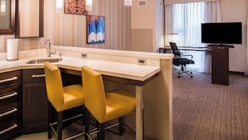 Une chambre d’hôtel équipée d’une kitchenette, de deux chaises jaunes, d’un bureau avec une chaise, d’un téléviseur à écran plat et d’une grande fenêtre avec des rideaux.