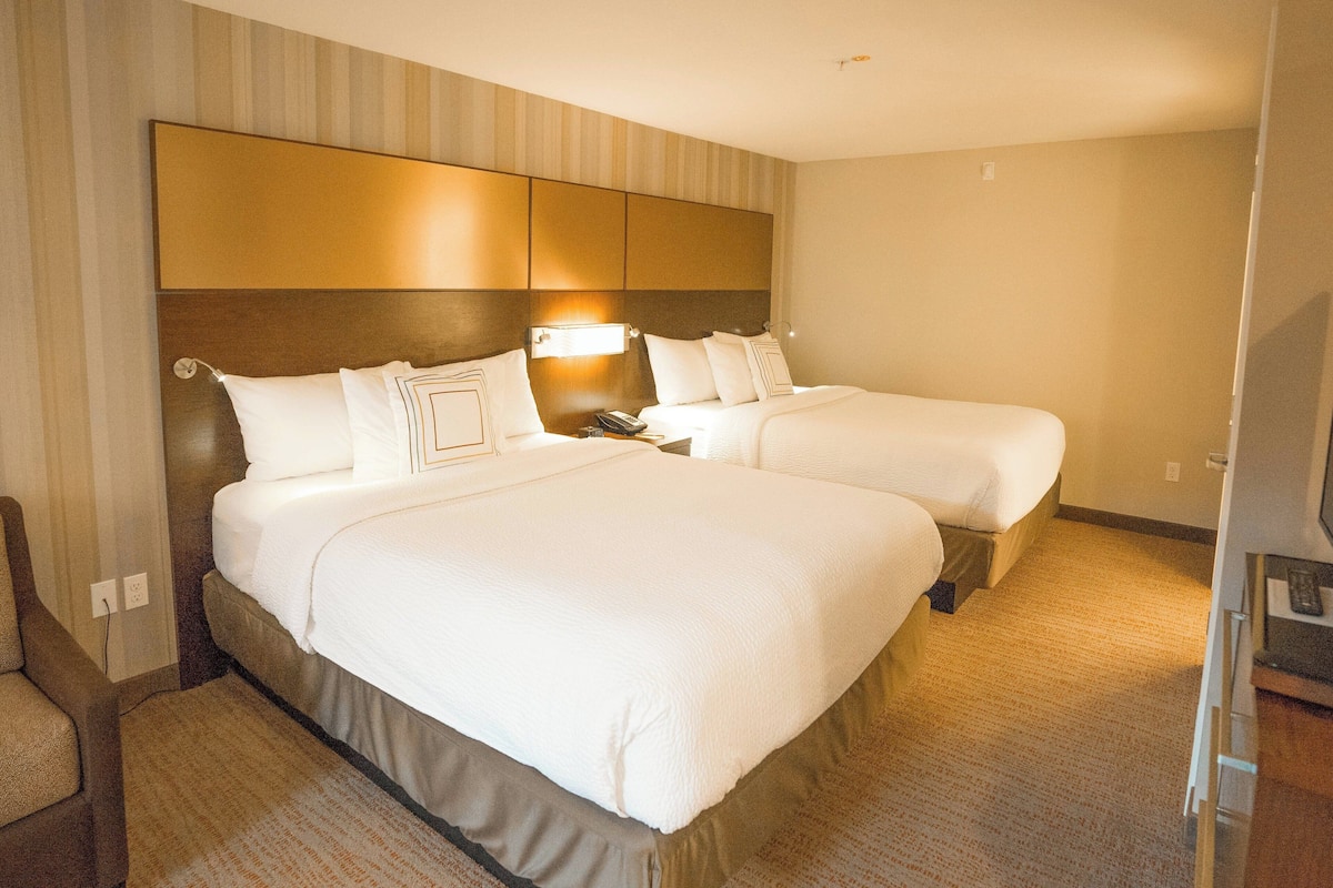 Ein Hotelzimmer mit zwei Betten, einem Kopfteil, Nachttischlampen und einem Teppichboden.