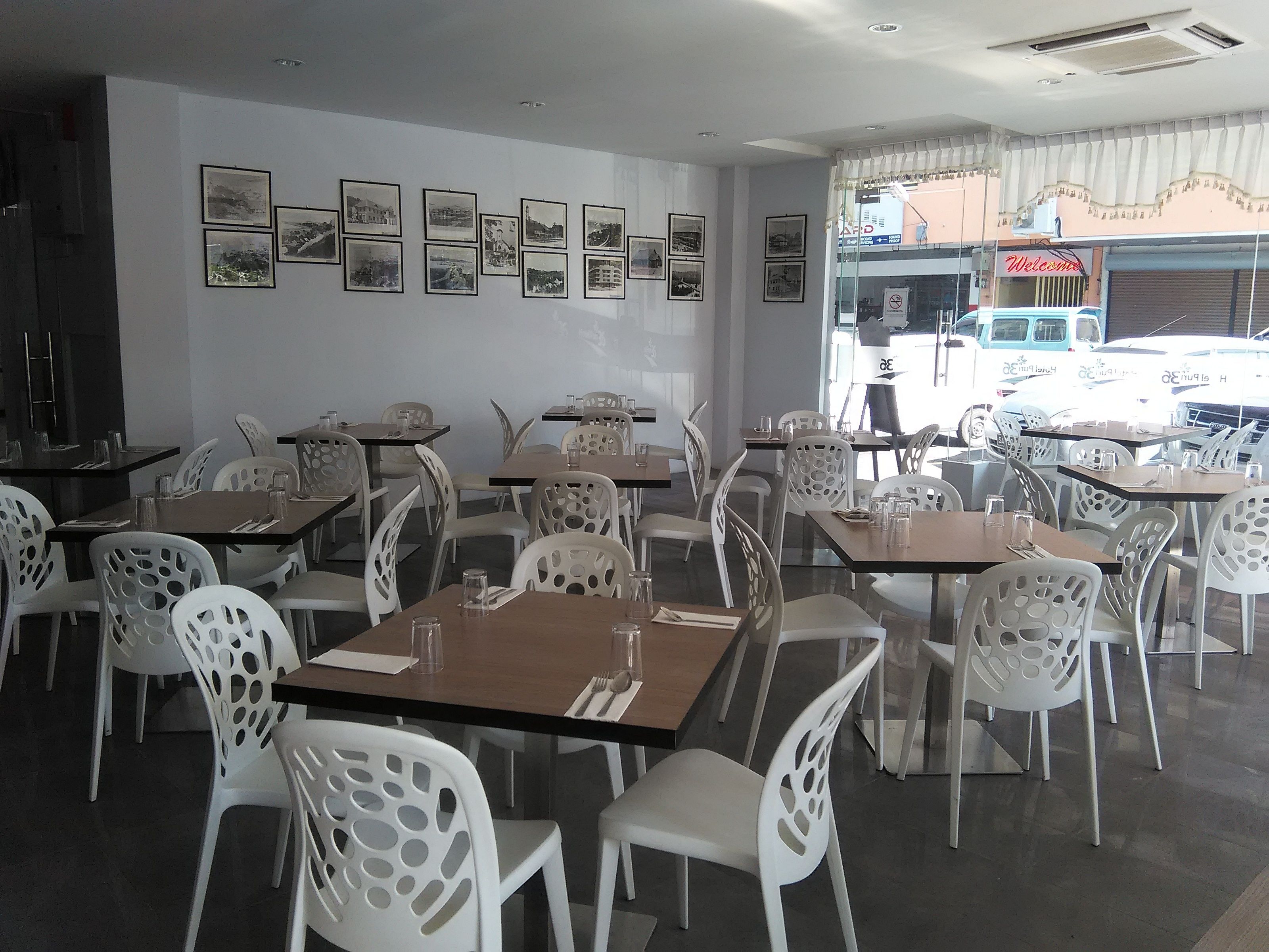 Un restaurante con sillas y mesas blancas, cuadros en la pared y vistas a vehículos estacionados afuera.