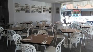 Un restaurante con sillas y mesas blancas, cuadros en la pared y vistas a vehículos estacionados afuera.