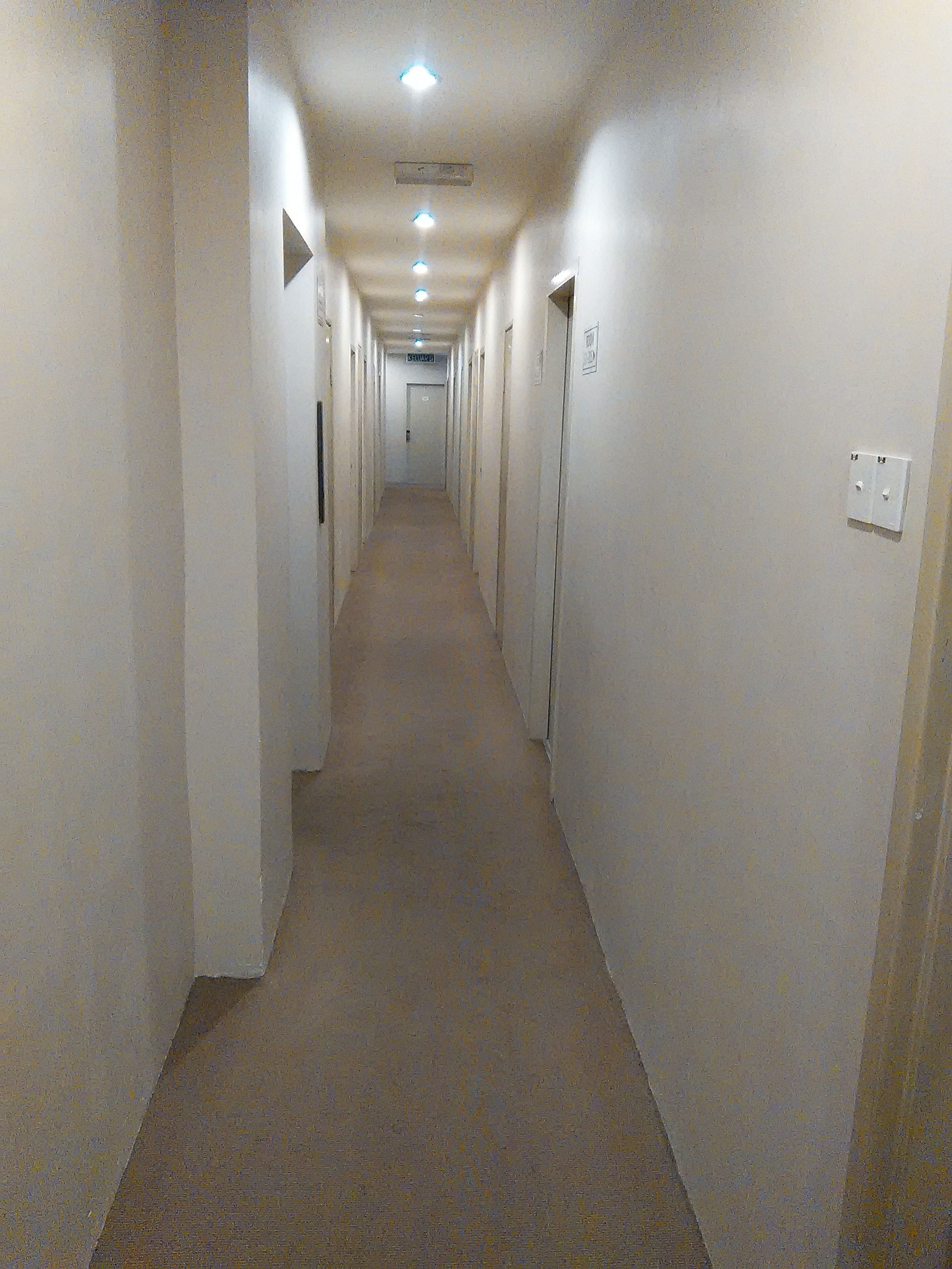 A long, narrow hotel corridor with doors on both sides, a carpeted floor, and a ceiling with recessed lighting.