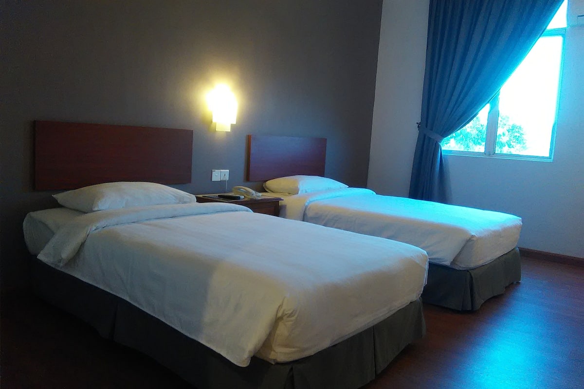A hotel room with two beds, a nightstand, a lamp, and a window with curtains.