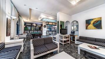 A modern living room with a sofa, a coffee table, and a TV. There is a bar area in the background with a pool table and a refrigerator.