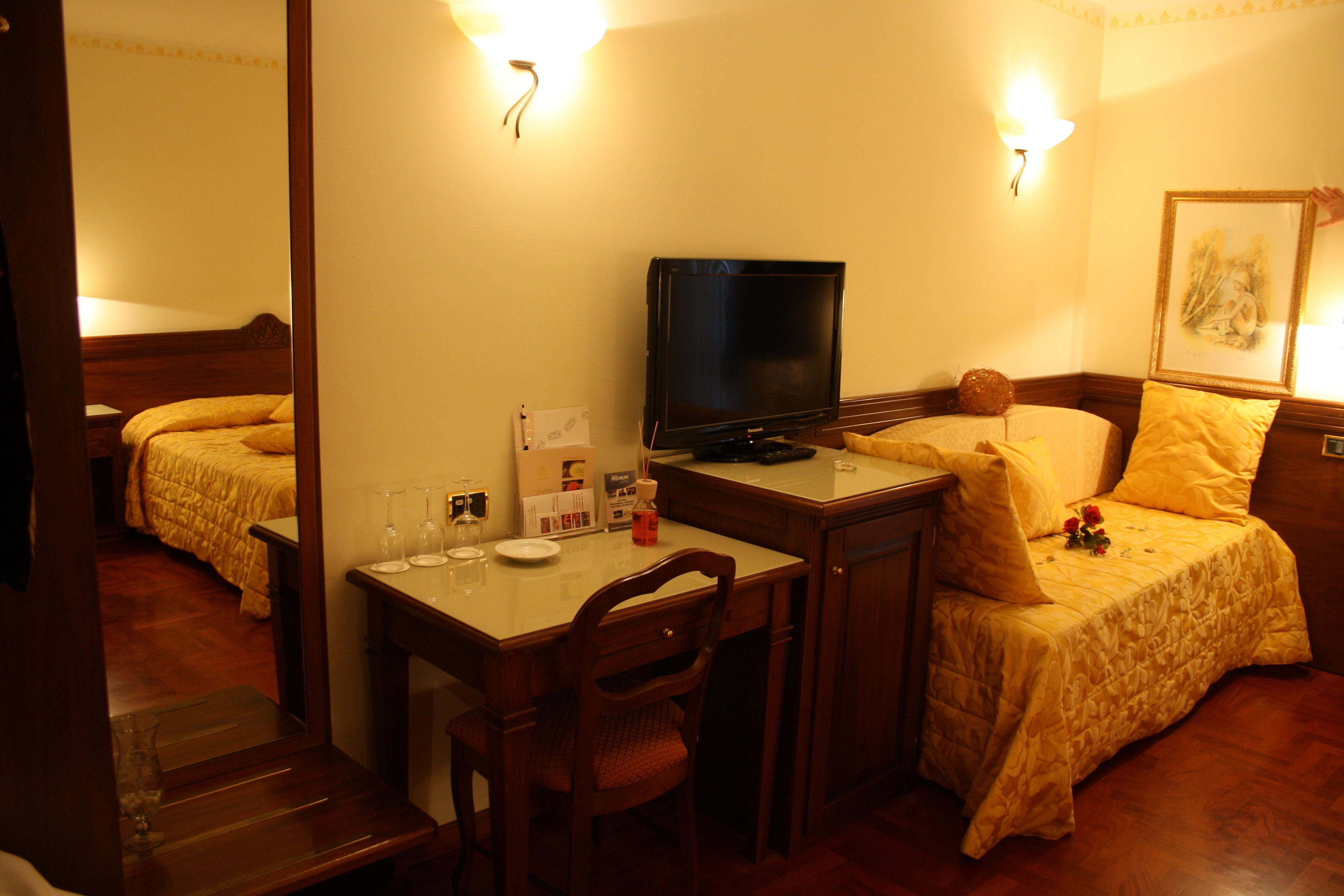 A hotel room with a bed, a desk with a chair, a television, and a bedside table with a lamp.