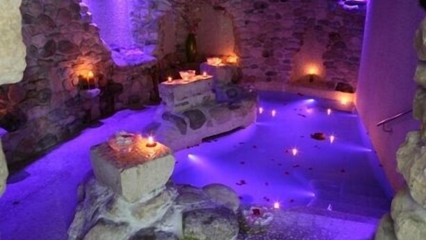 Una habitación de paredes de piedra con una piscina central, velas y un banco de piedra.