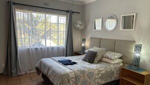 A bedroom with a bed, a nightstand, a bedside table, a window with curtains, a wall-mounted fan, and a mirror.
