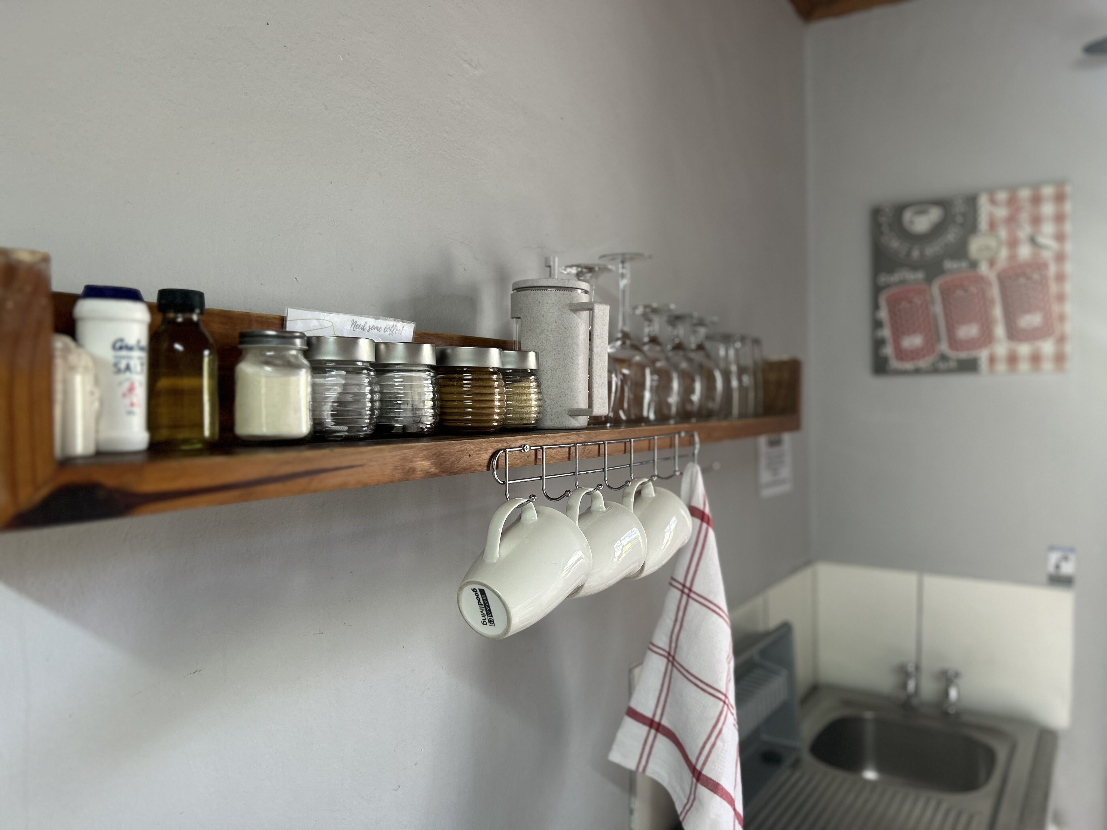 A kitchen shelf with various jars and containers, a hanging rack with mugs, and a kitchen sink.