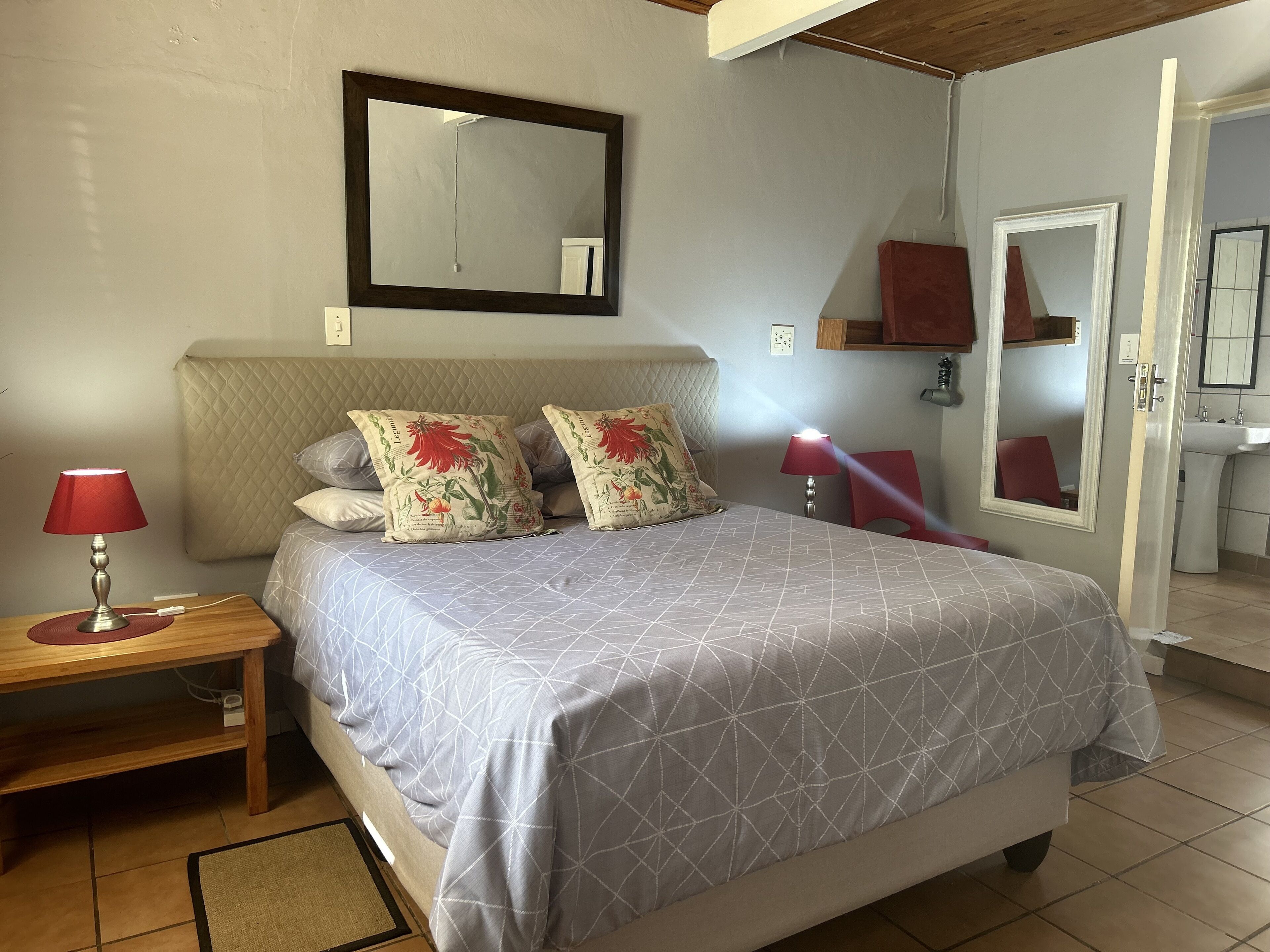 A bedroom with a bed, two bedside lamps, a nightstand, a dresser, a mirror, and a bathroom visible through an open door.