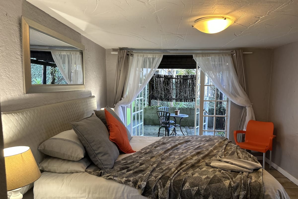 A bedroom with a bed, a nightstand, a window with curtains, a chair, and a view of the outside.