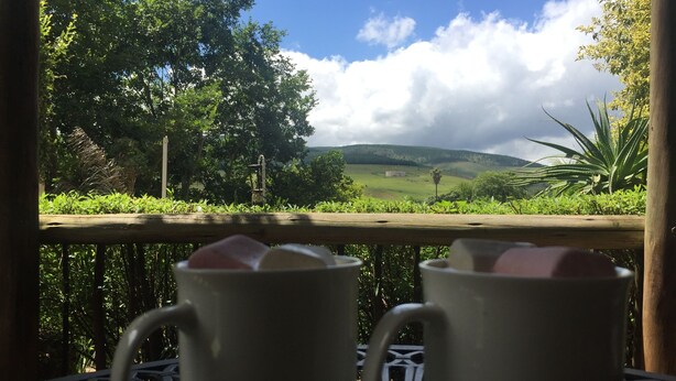 Two white mugs on a metal table with a view of a green landscape and hills in the background.