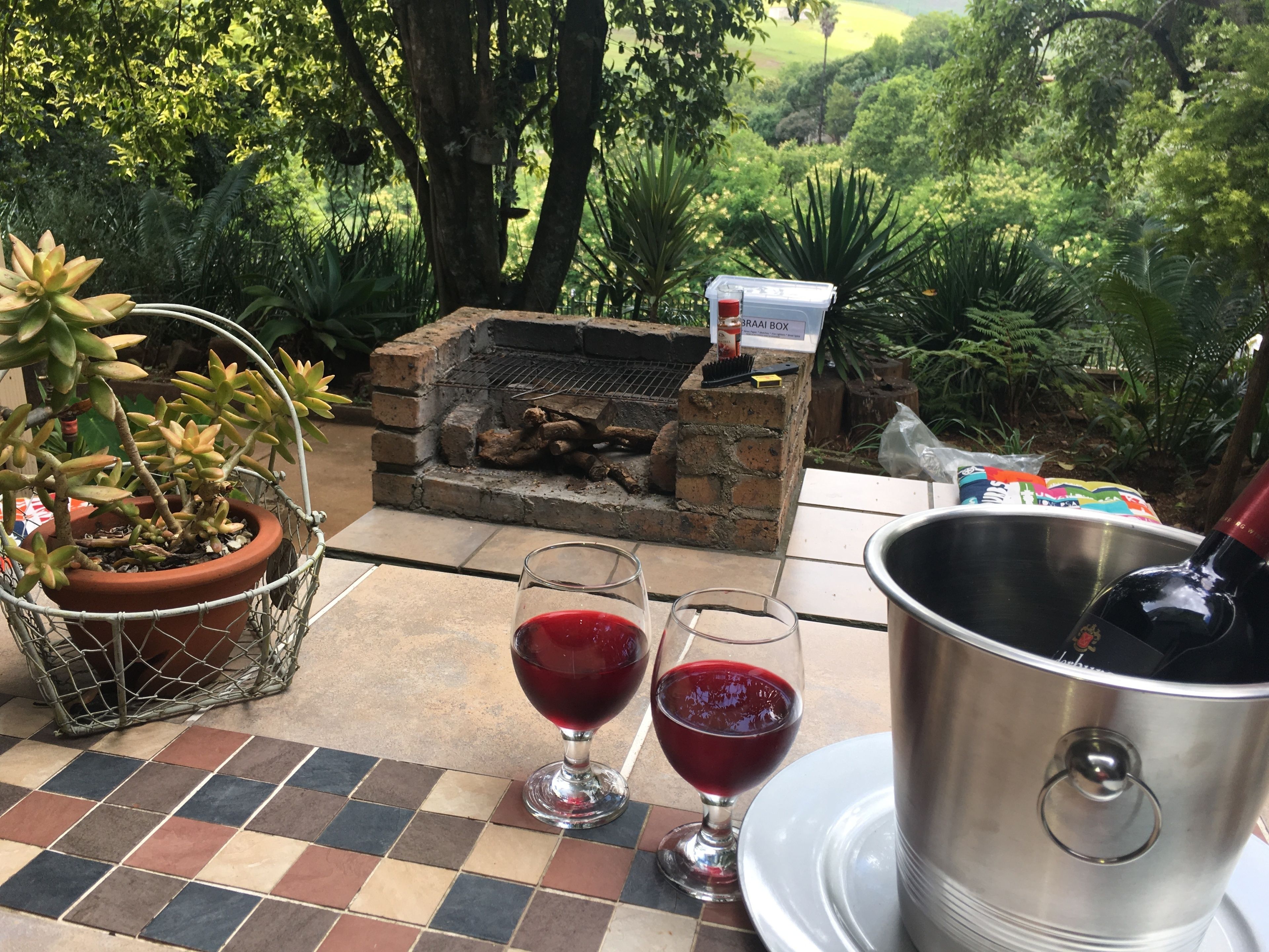 Un patio exterior con un brasero de ladrillo, dos copas de vino, un cubo de metal y una planta en maceta.