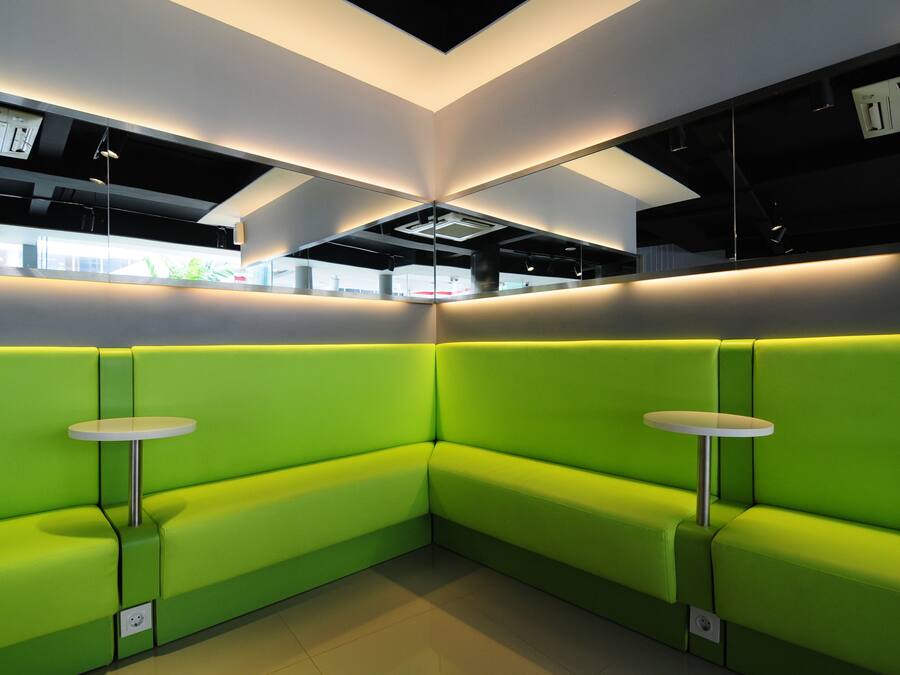 A modern interior with green seating and small tables, featuring a geometric ceiling design and visible air conditioning units.