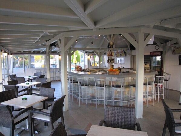 Ein Straßencafé mit Weißlchen Stühhlen und Tischen, einer Bar in der Mitte und einem überdachten Sitzbereich.