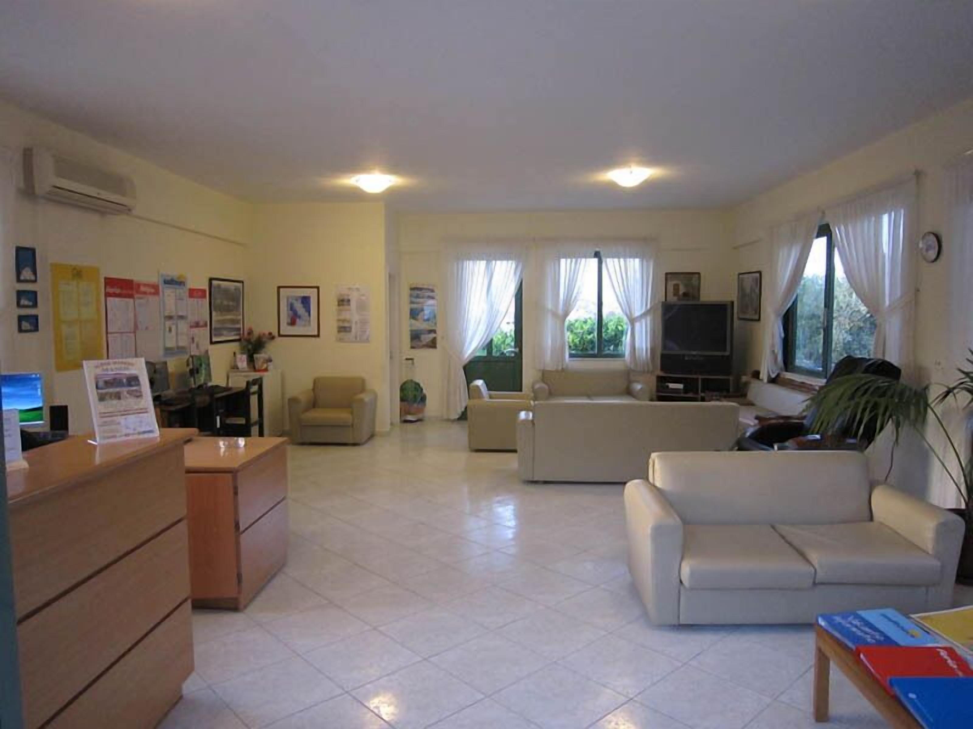 A spacious living room with a sofa, armchairs, and a TV. There is a wooden desk with a computer and a chair. The room has large windows with curtains and a ceiling light.