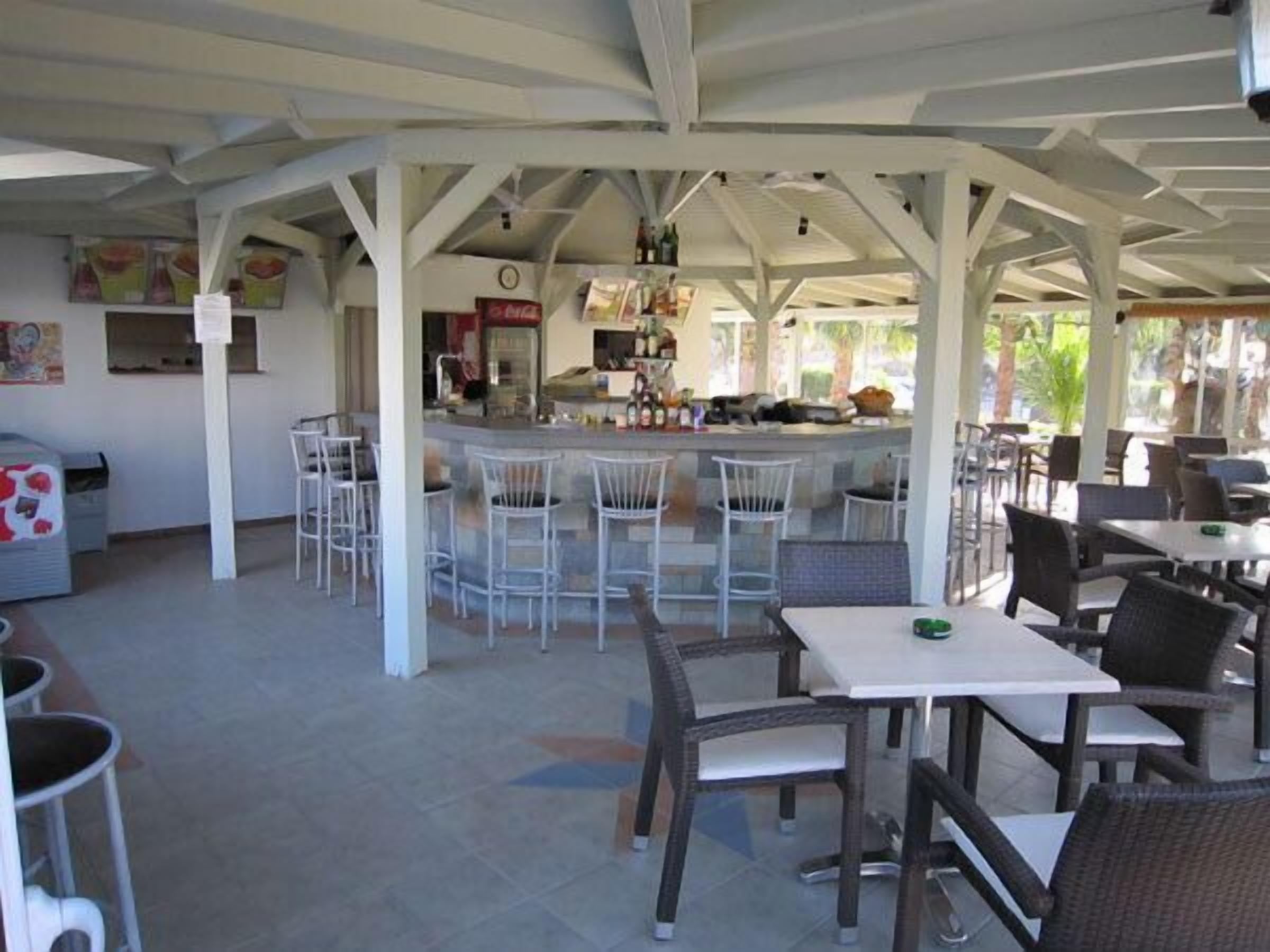 Un café al aire libre con taburetes y mesas blancos, una zona de bar con botellas y un cubo de basura.