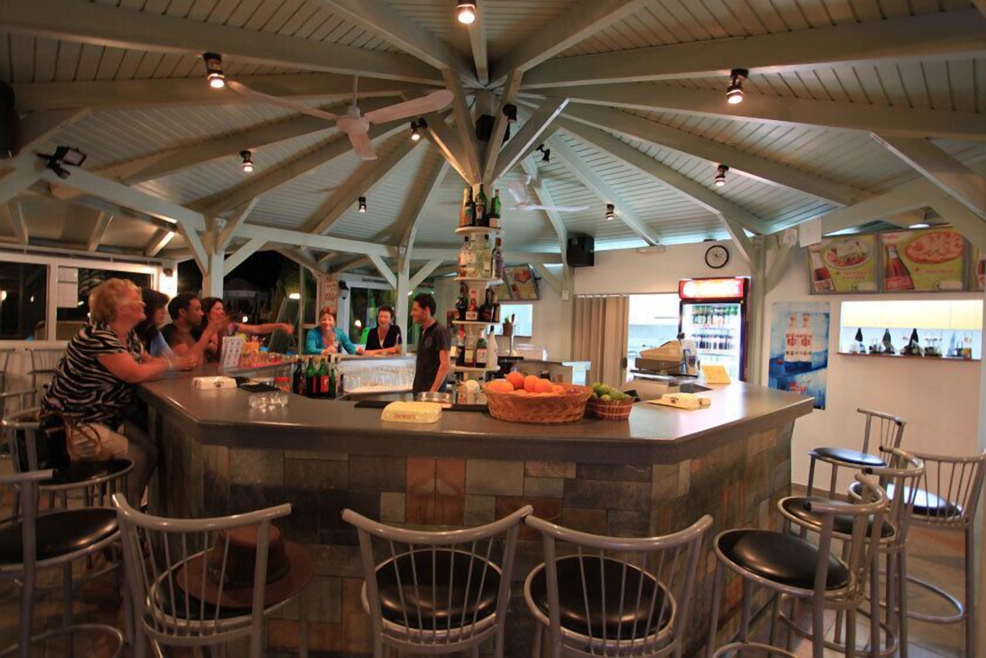 Un espace bar doté d’un plafond haut et de poutres apparentes, avec un comptoir central, des tabourets, ainsi qu’un assortiment de bouteilles et de collations.