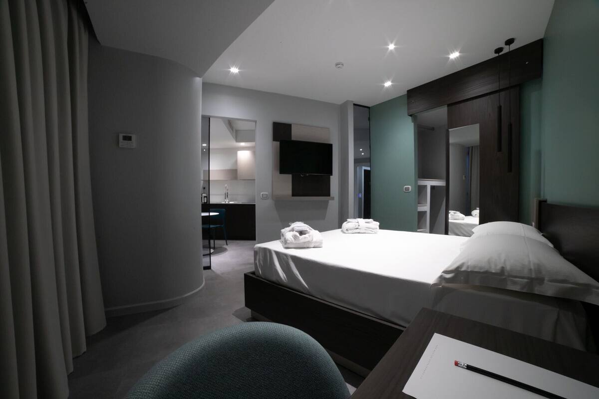 A modern hotel room with a bed, a desk, a chair, a TV, and a bathroom visible through an open door.