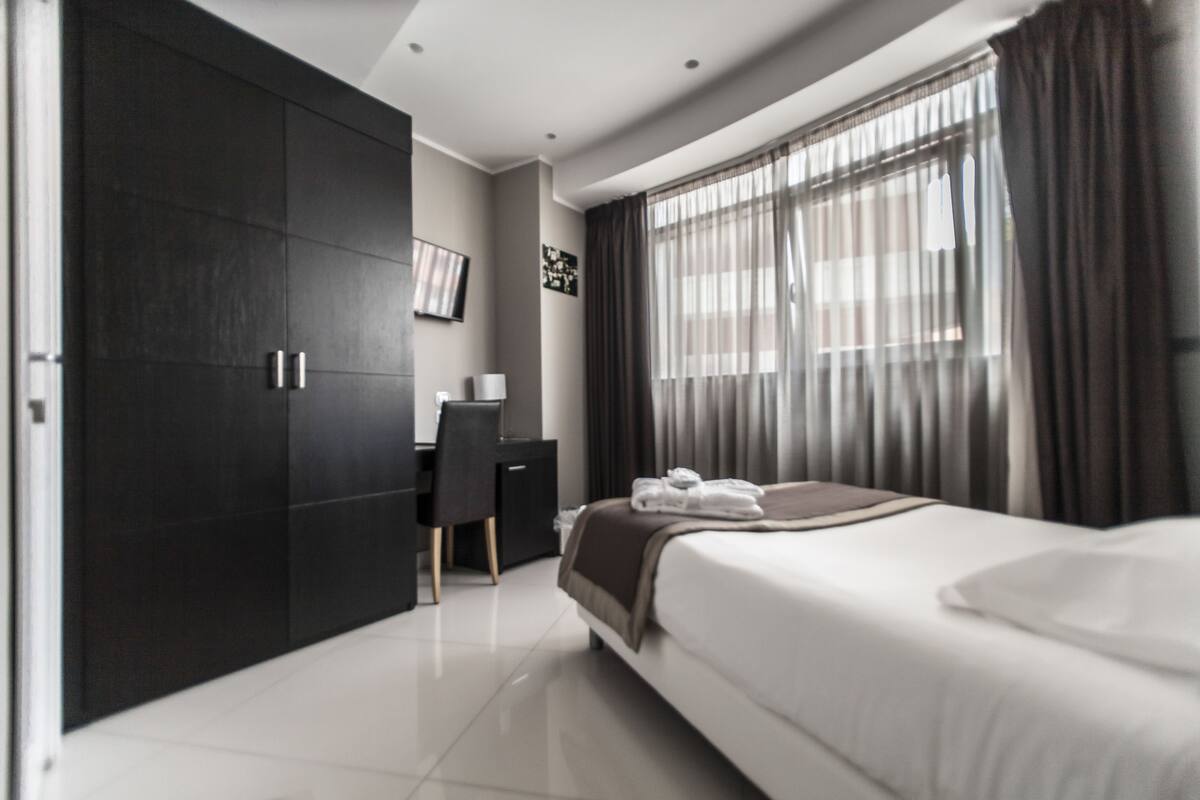 A modern hotel room with a large bed, a desk, and a wardrobe.