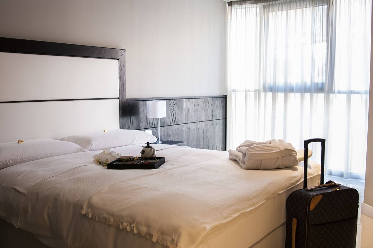 A neatly made bed with a tray of tea and flowers, a suitcase, and a lamp on a nightstand.