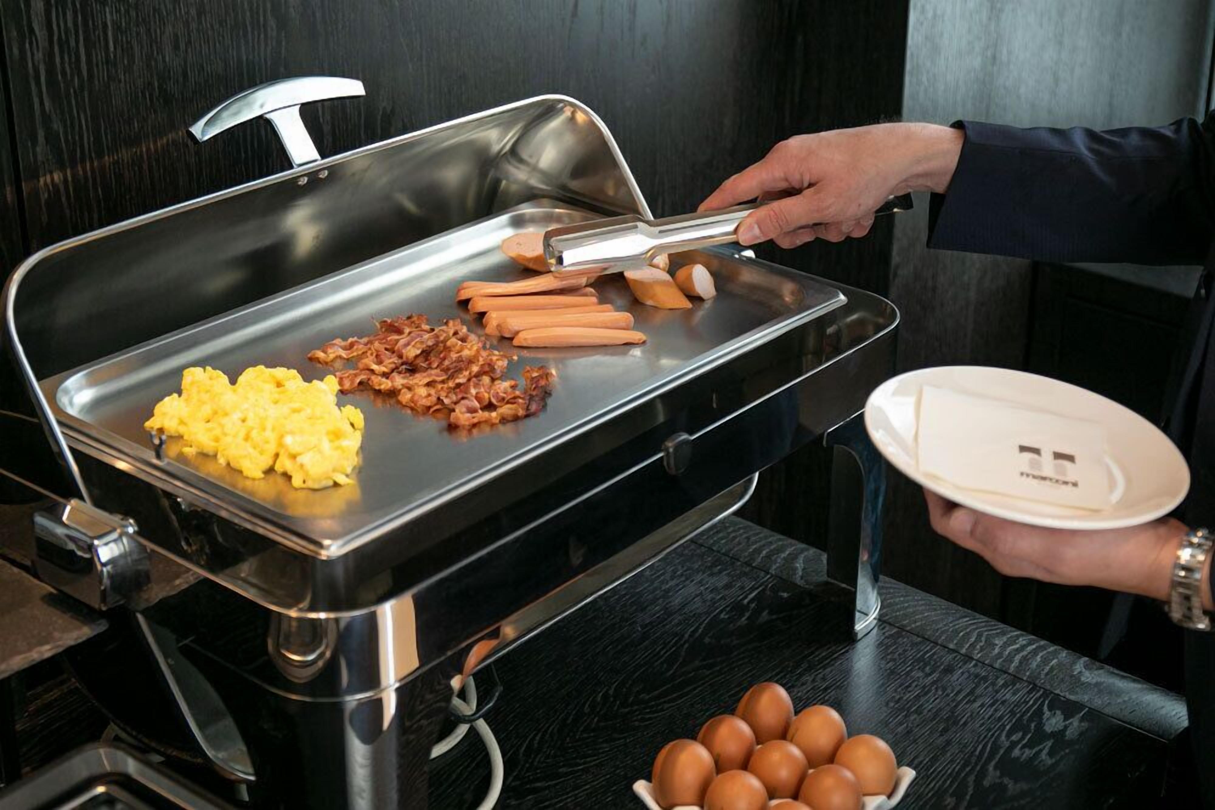 A buffet setup with scrambled eggs, bacon, and sausages on a grill, and a hand holding a plate with a napkin.