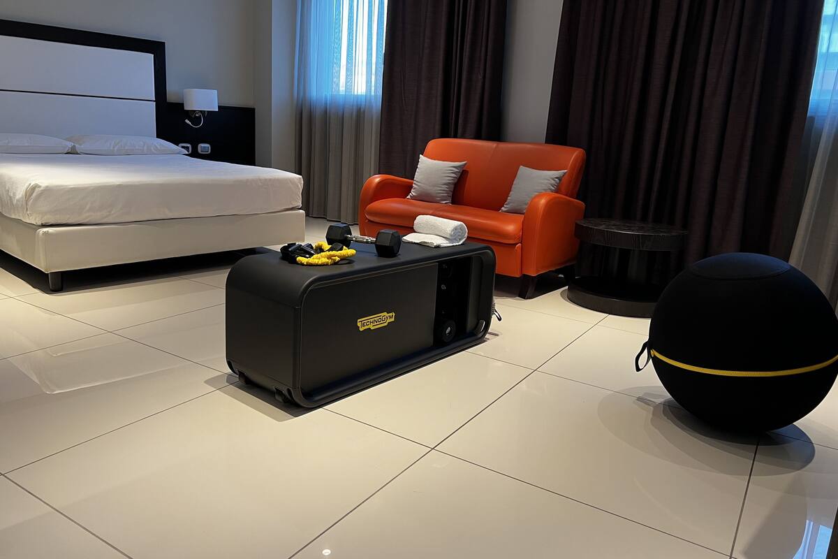 A hotel room with a bed, a red armchair, a black suitcase, and a black exercise ball.