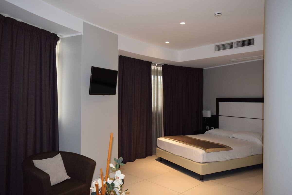 A hotel room with a bed, a chair, a TV mounted on the wall, and a window with curtains.