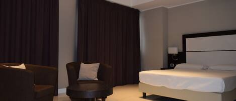 A modern hotel room with a bed, two armchairs, a small round table, and a nightstand with a lamp.