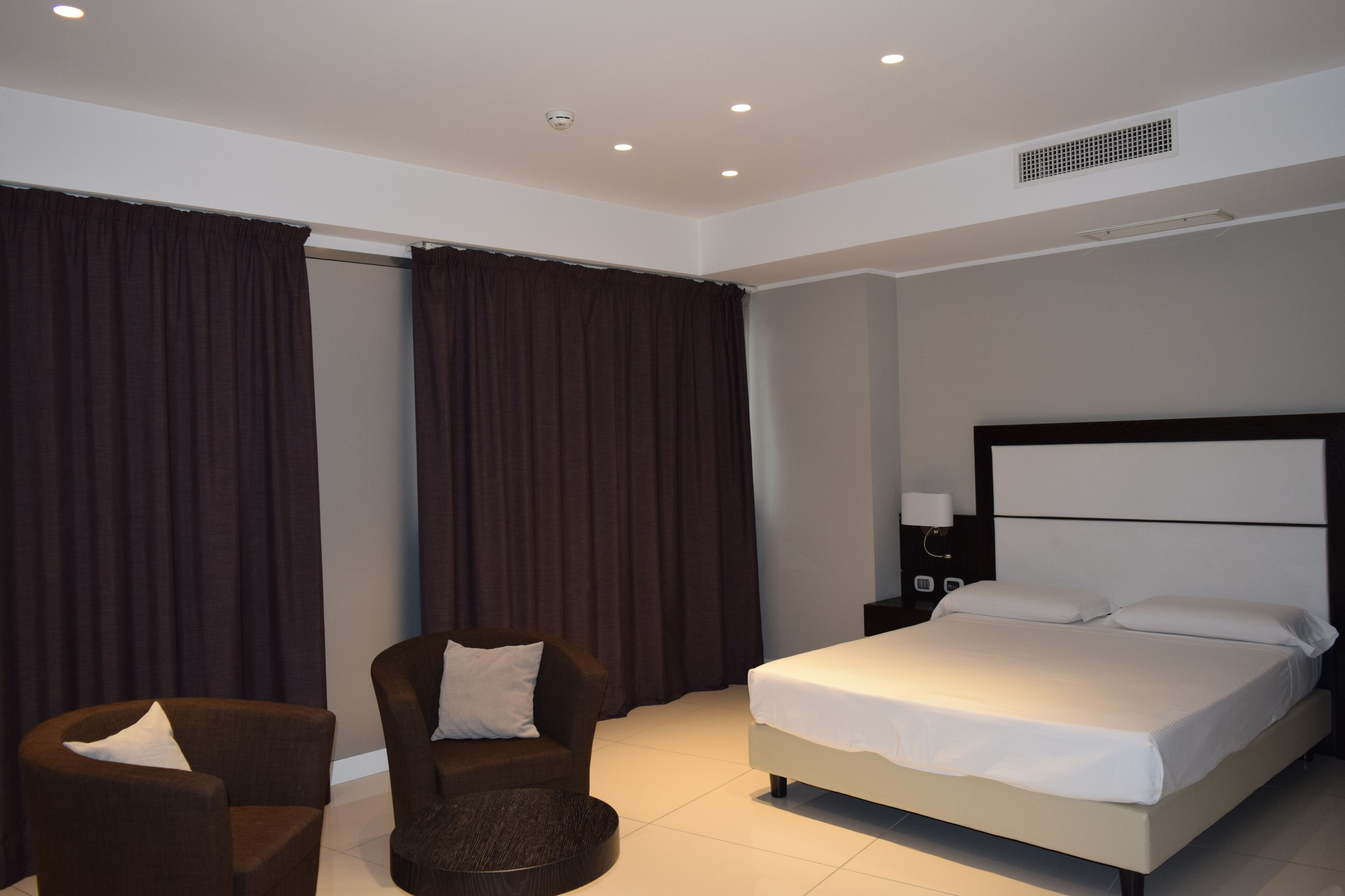 A hotel room with a bed, a chair, a nightstand, and a large window with curtains.