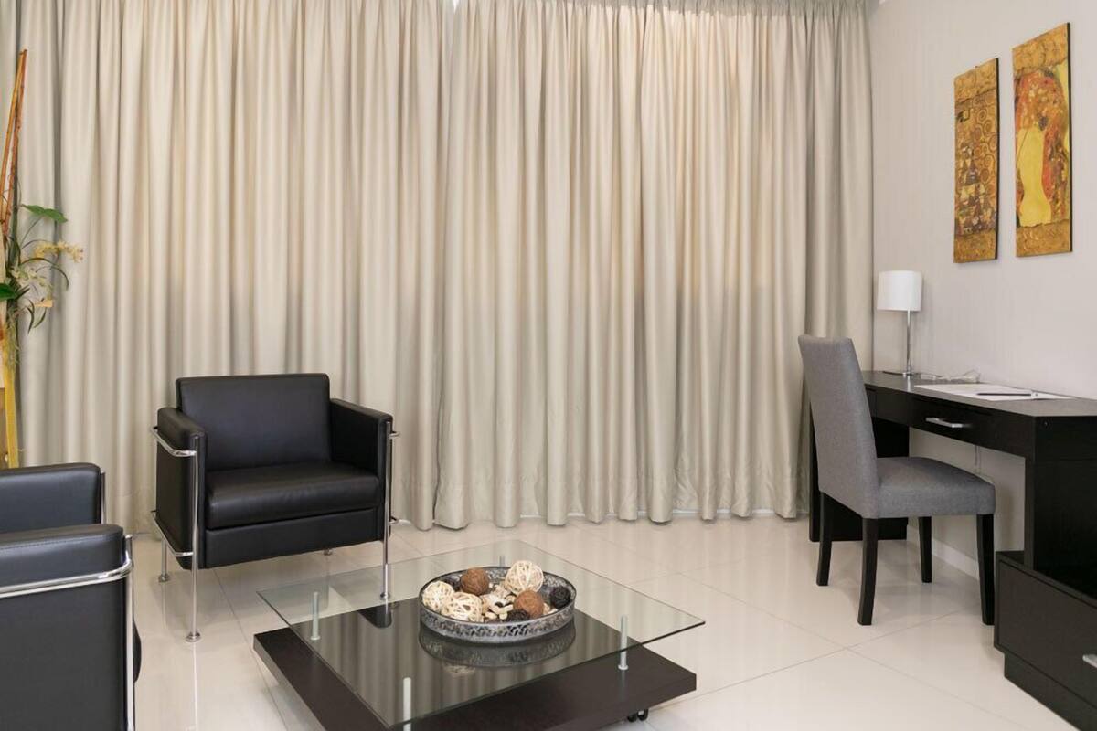 A modern living room with a glass coffee table, a black sofa, a grey office chair, and a desk with a lamp.